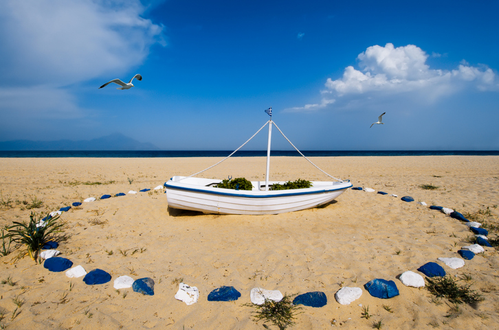 Ein weißes, landgestütztes Boot umgeben von einem Kreis aus blau-weißen Steinen an einem weiten Sandstrand in Sarti.