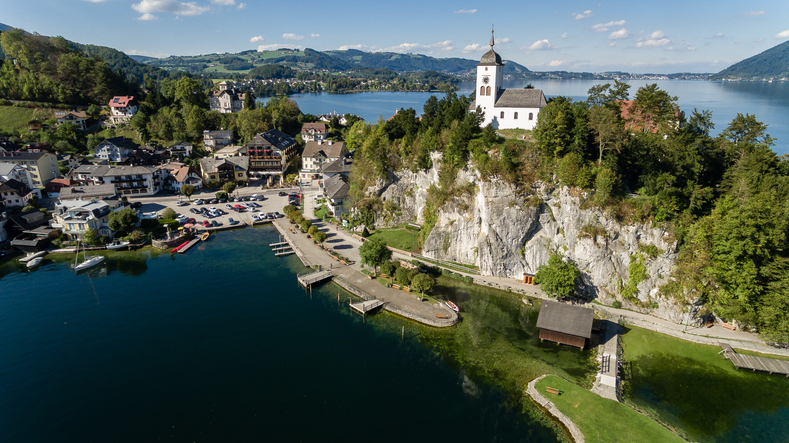 Eine kleine, weiße Kirche thront auf einem markanten Felsen direkt an einem tiefblauen See, umgeben von grüner Natur.