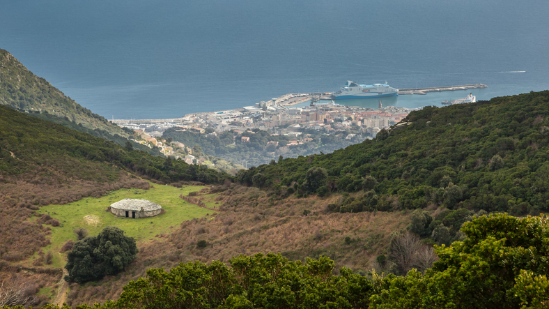 Ein verlassenes, kreisrundes Steingebäude steht einsam auf einer grünen Wiese in einer hügeligen Landschaft mit Blick auf den Hafen und das Meer im Hintergrund.