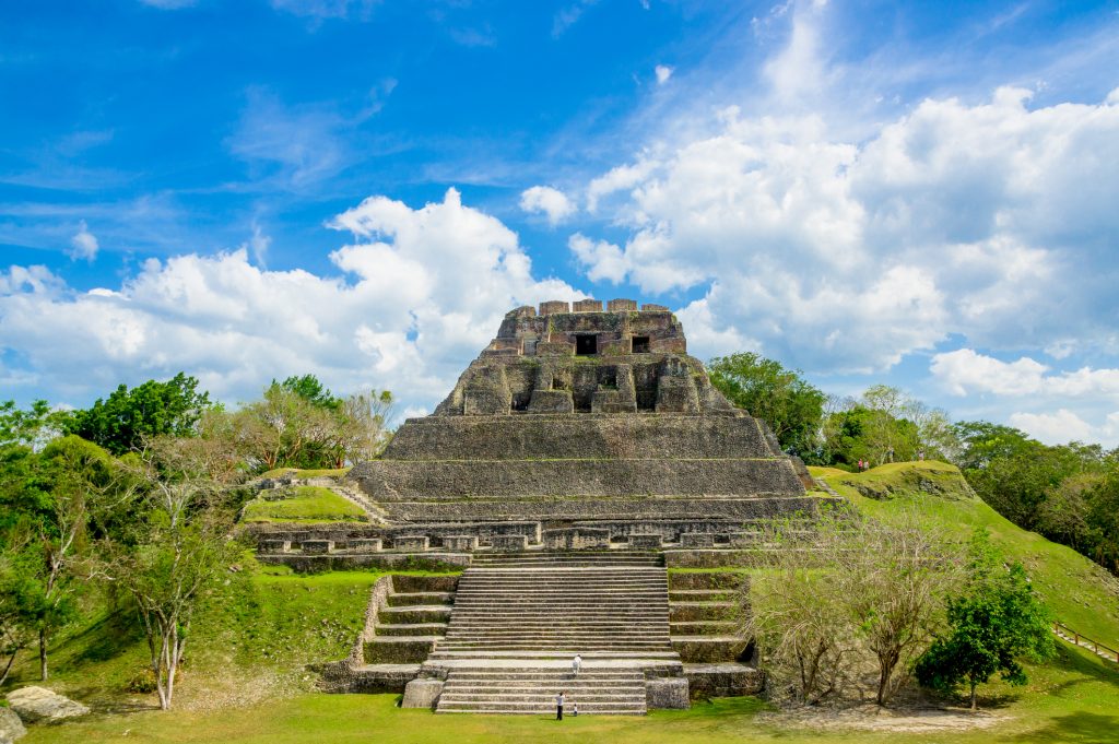 Eine massive, stufenförmige Maya-Tempelanlage ragt majestätisch vor einem strahlend blauen Himmel mit weißen Wolken auf.