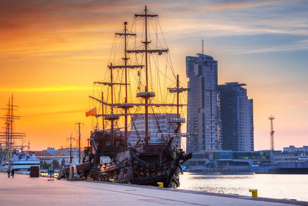 Ein majestätisches Segelschiff mit vollen Masten vor dem goldenen Licht des Sonnenuntergangs und den modernen Bürogebäuden von Gdynia.
