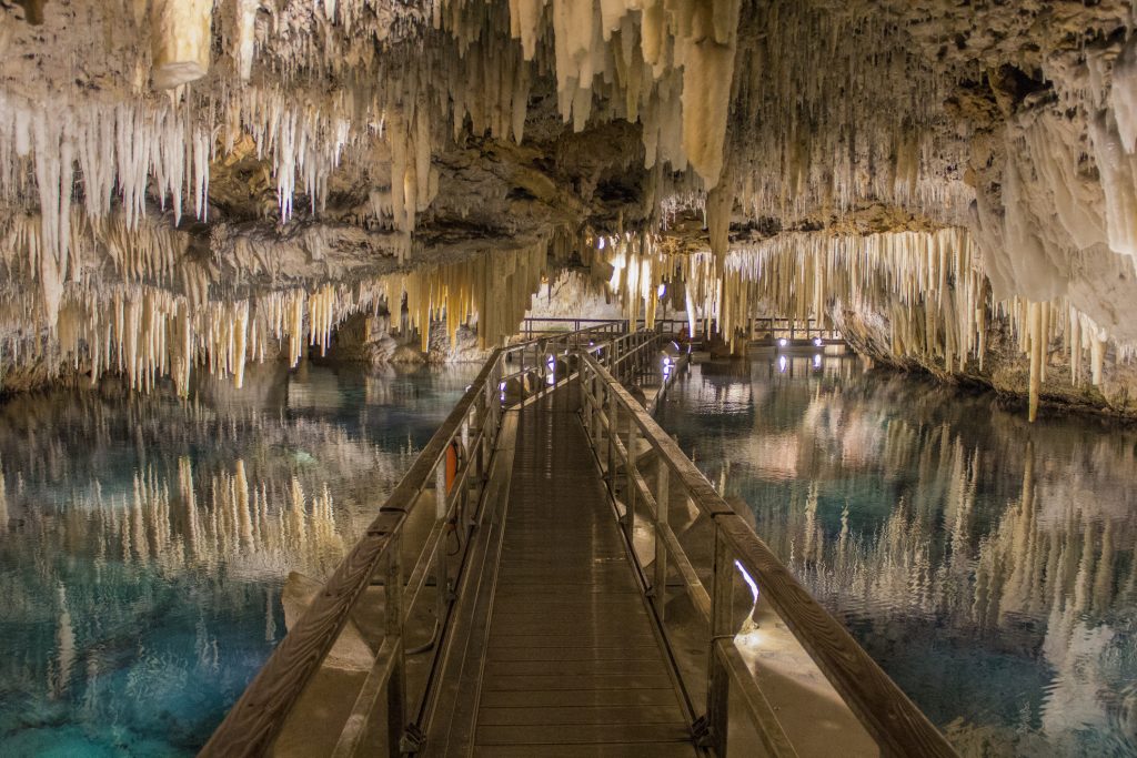 Ein Holzsteg führt durch eine unterirdische Höhle mit zahlreichen Stalaktiten über einem spiegelglatten See.