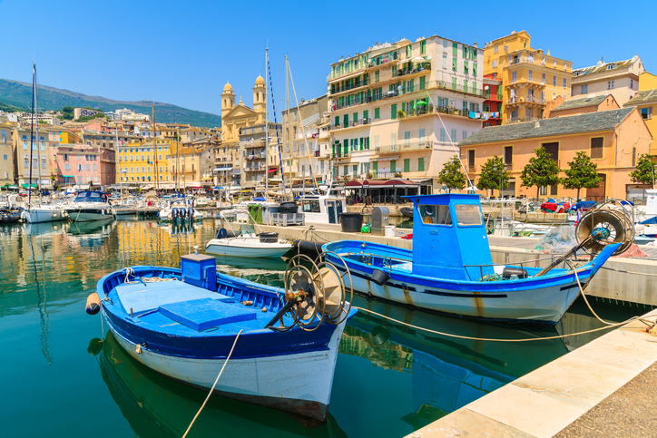 Blaue Fischerboote liegen friedlich im Wasser des Hafens von Bastia vor den hohen, bunten Häuserfronten.