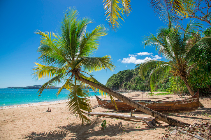 Eine Palme neigt sich über einen goldenen Sandstrand, an dem ein altes hölzernes Auslegerboot im klaren türkisfarbenen Wasser liegt.