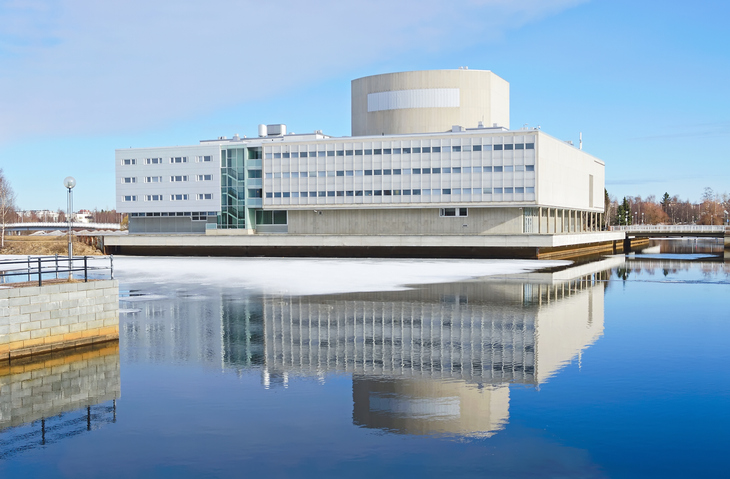 Ein modernes, helles Gebäude am Wasser, dessen Fassade sich im ruhigen Flussbett spiegelt.