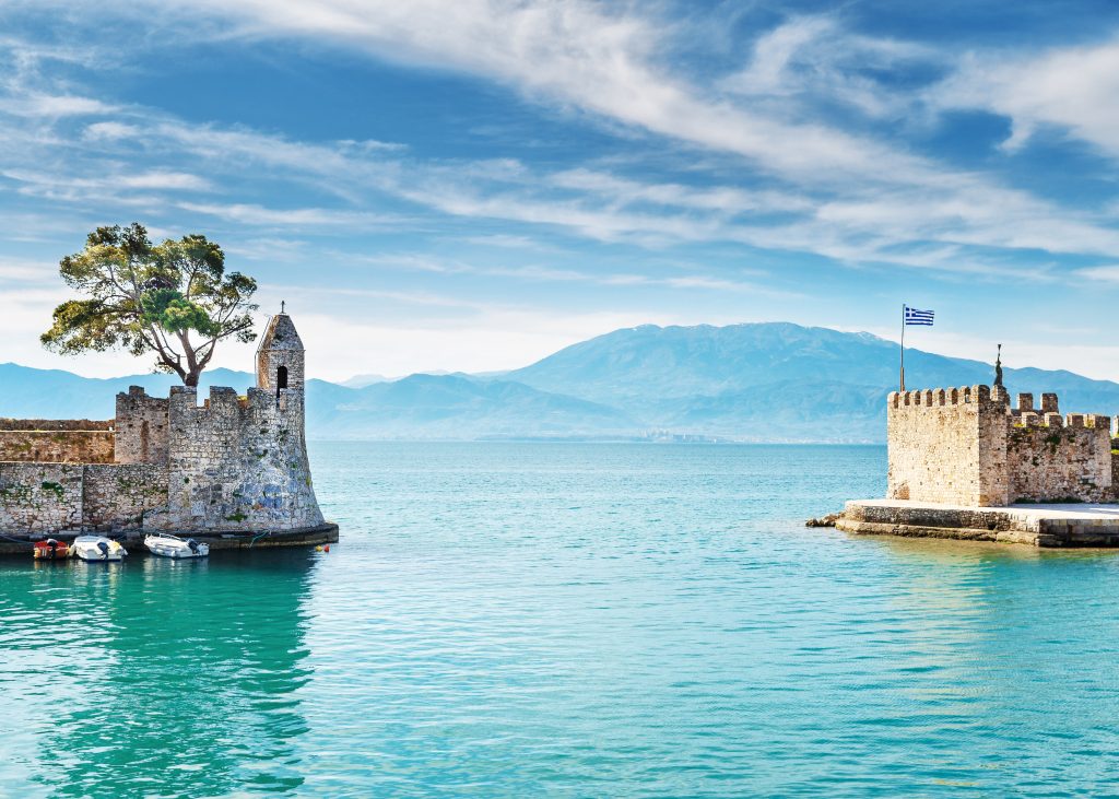 Zwei historische Steinbastionen flankieren die Hafeneinfahrt mit blauem Wasser und Bergen im Hintergrund.