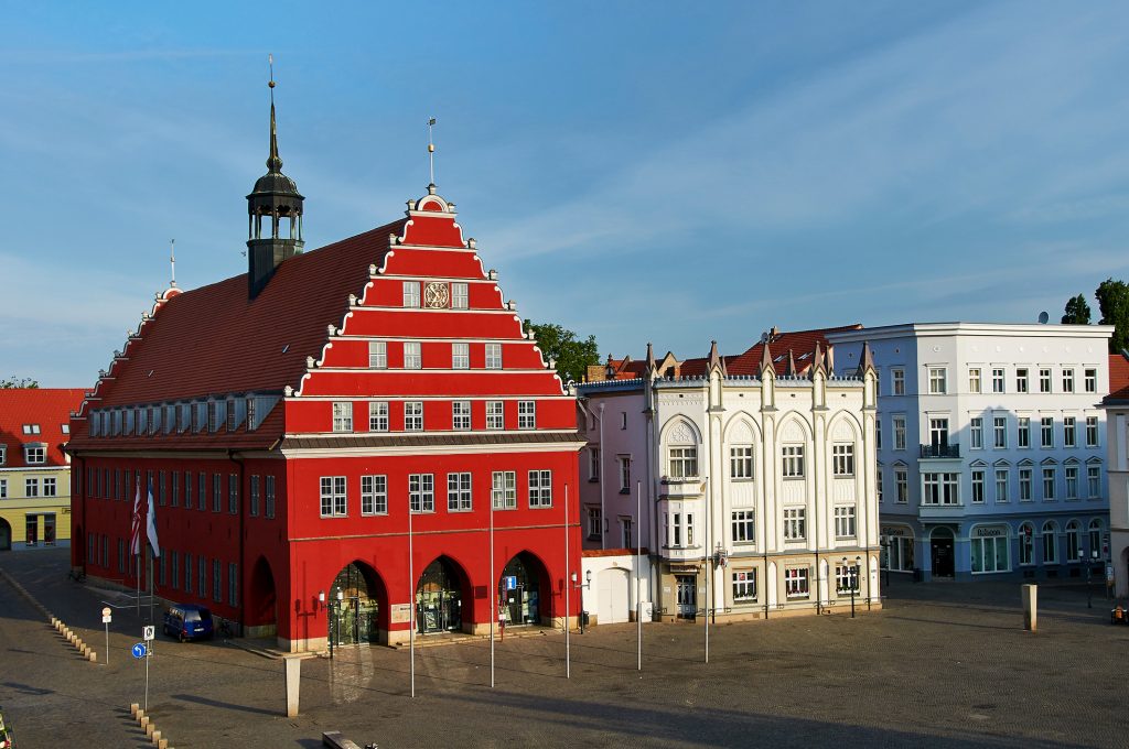 Das markante, leuchtend rote Rathaus von Greifswald steht an einem weitläufigen, gepflasterten Marktplatz.