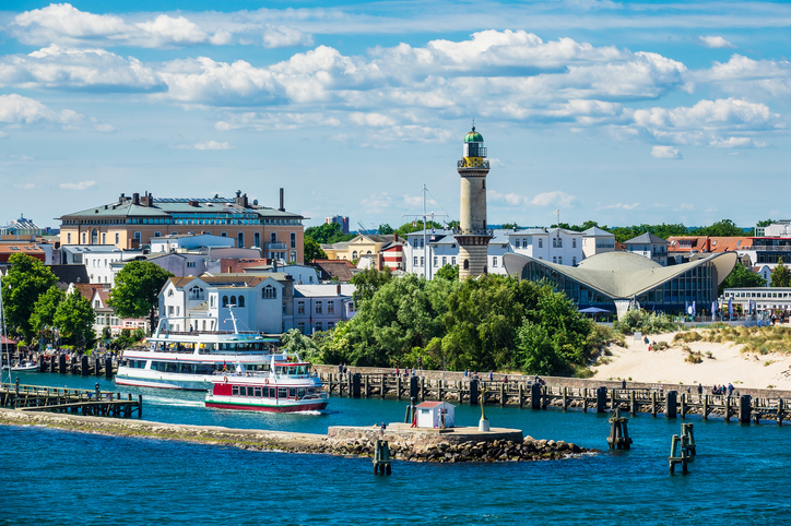 Ein sommerlicher Blick auf die Hafeneinfahrt von Warnemünde mit Ausflugsschiffen, Leuchtturm und Strandbereich.
