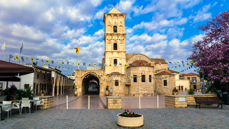 Historische Kirche mit Vorplatz, Arkadengang und Frühlingsbaum unter wolkigem Himmel in Larnaka