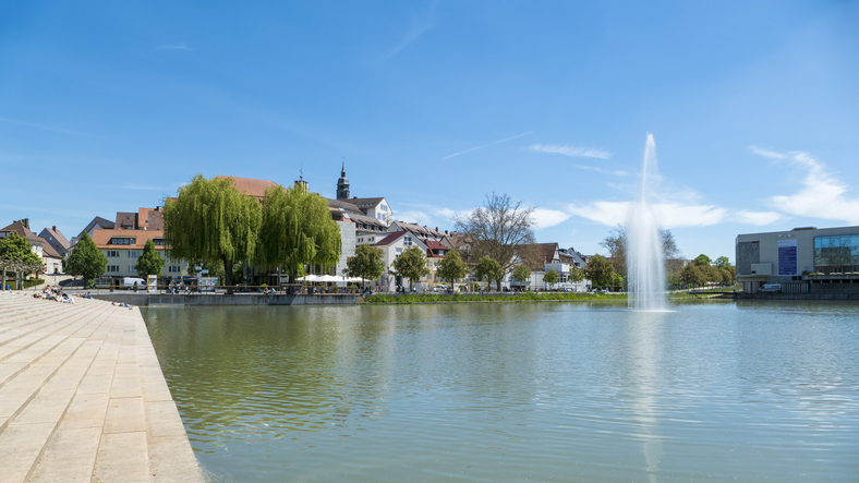 See in Böblingen mit Wasserfontäne, Uferpromenade und Stadtgebäuden im Hintergrund.