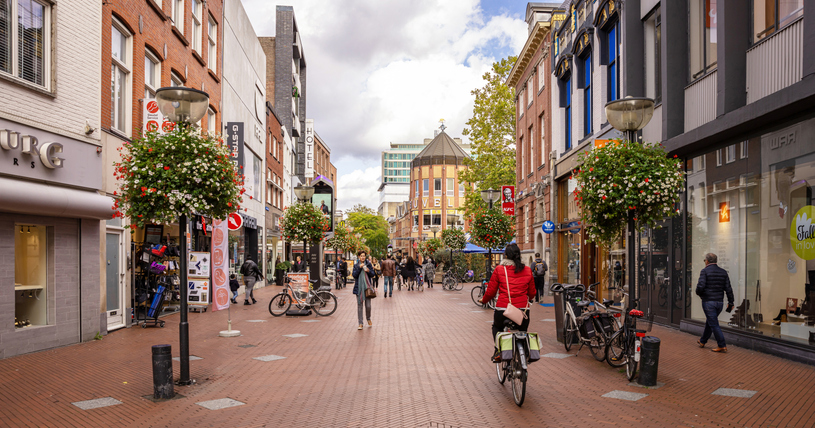 Belebte Einkaufsstraße in Eindhoven mit Menschen, Fahrrädern und Blumenampeln.
