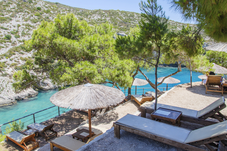 Sonnenliegen und Strohdächer auf einer Terrasse über einer kleinen türkisfarbenen Bucht auf Zakinthos