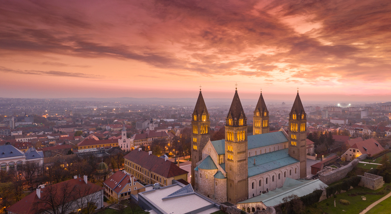 Kathedrale von Pécs bei Sonnenuntergang mit leuchtendem Himmel, vier Türmen und Stadtpanorama.