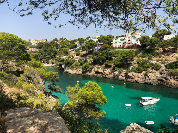 Schmale türkisfarbene Meeresbucht von Cala Pi mit Booten, Felsen, Pinien und Häusern im Hintergrund.
