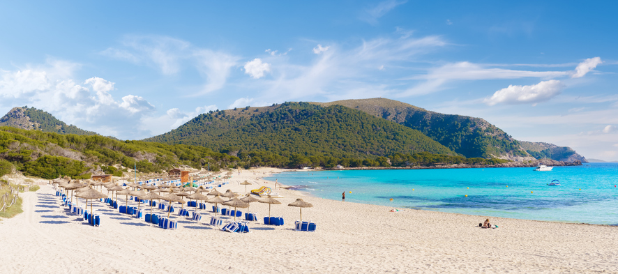 Weiter Sandstrand der Cala Agulla mit Liegen, Sonnenschirmen, Bergen und türkisblauem Meer.