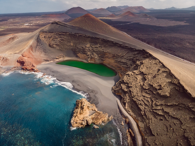 Aus der Vogelperspektive sind Küste, Kraterlandschaft und eine grüne Lagune auf Lanzarote zu sehen.