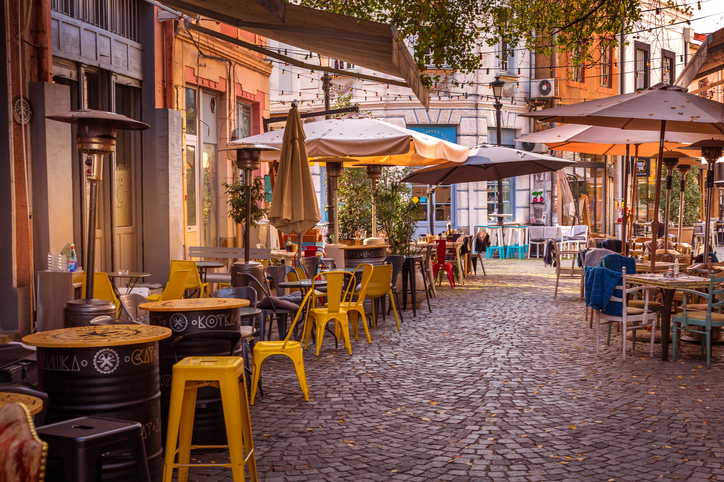 Belebte Kopfsteinpflastergasse in der Altstadt von Plowdiw mit Cafés, Tischen, Sonnenschirmen und bunten Stühlen.