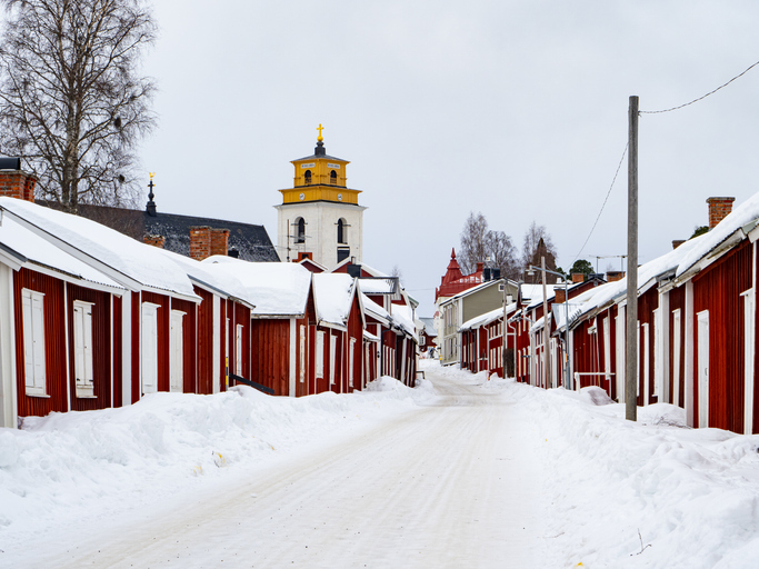Verschneite Straße mit roten Holzhäusern und Kirche im Hintergrund in Gammelstad bei Luleå.