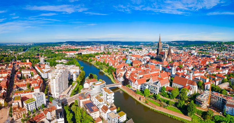 Weite Luftaufnahme von Ulm mit Flusslauf, Brücken, Altstadt und dem Ulmer Münster unter blauem Himmel.
