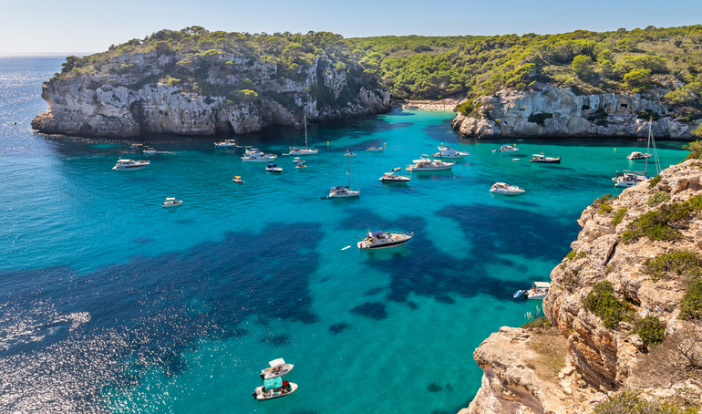 Eine türkisfarbene Bucht auf Menorca mit vielen kleinen Booten, Felsen und grüner Küstenlandschaft.