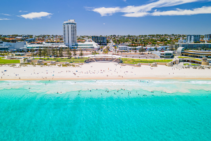 Luftaufnahme eines hellen Sandstrands in Perth mit türkisblauem Meer, Badenden und Stadtbebauung im Hintergrund.