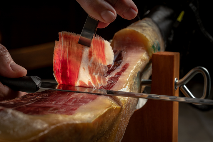 Eine Hand schneidet hauchdünne Scheiben Jamón an einem Schinkenhalter