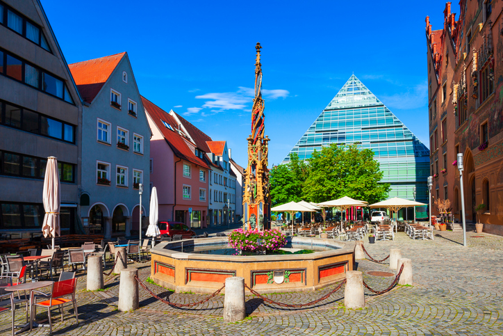 Platz in Ulm mit Brunnen, buntem Zierbrunnen und gläserner pyramidenförmiger Architektur im Hintergrund.