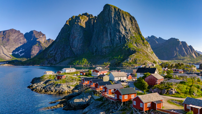 Kleine rote Häuser liegen an einer felsigen Küste vor einem steilen Berg am Meer in Norwegen.