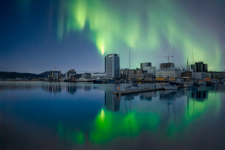 Grüne Nordlichter leuchten über dem Wasser und einer Stadt mit Booten in Norwegen.