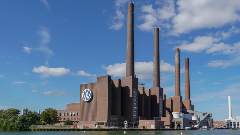 Das Volkswagenwerk in Wolfsburg mit VW-Logo, Industriegebäude und hohen Schornsteinen am Wasser.