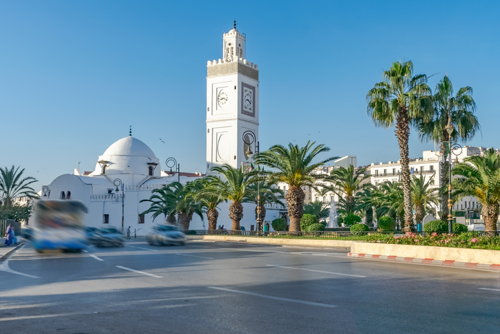 Eine weiße Moschee mit Kuppel, Minarett und Palmen steht an einer breiten Straße in Algier.