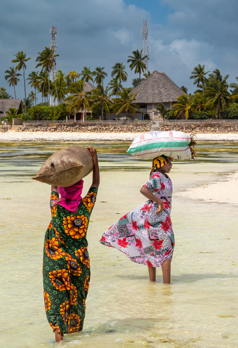 Zwei Frauen gehen im flachen Wasser an der Küste von Sansibar und tragen Waren.