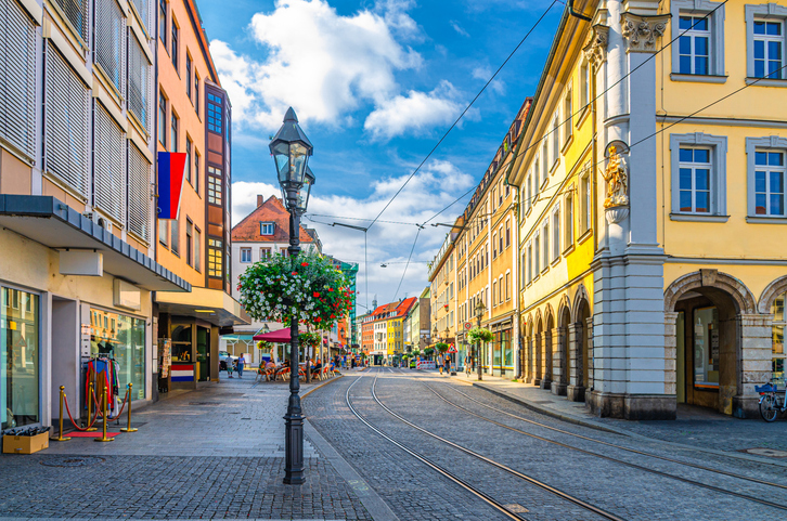 Eine Straße in der Würzburger Innenstadt mit historischen Fassaden, Straßenbahnschienen und einer Blumenlaterne.