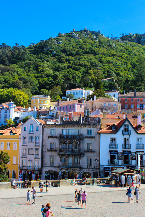 Auf einem Platz in Sintra stehen Besucher vor bunten Häusern, darüber liegt die Burg auf dem Hügel.