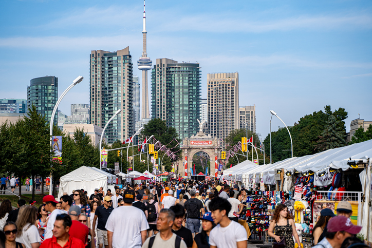 Menschenmenge auf einer breiten Straße in Toronto mit Marktständen und CN Tower im Hintergrund.