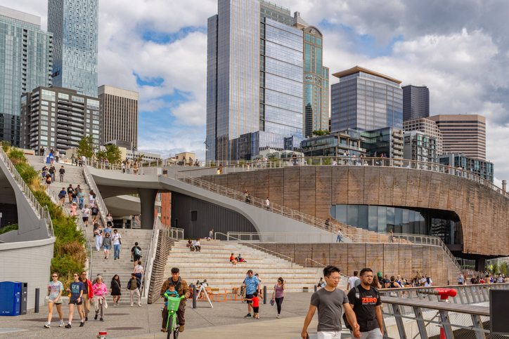 Menschen spazieren an der modernen Waterfront von Seattle zwischen Treppen, Holzfassaden und Hochhäusern.