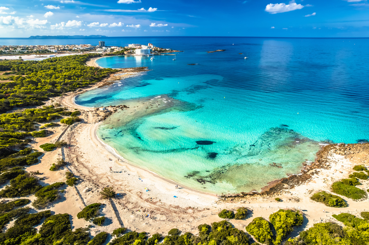 Luftaufnahme einer geschwungenen Bucht bei Es Trenc mit hellem Sand, türkisfarbenem Wasser und niedriger Vegetation.