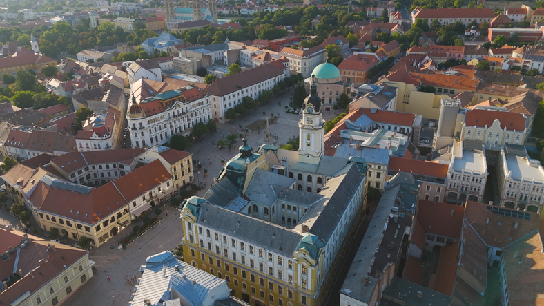 Luftaufnahme von Pécs mit historischen Gebäuden, roten Dächern, Plätzen und der grünen Kuppel im Stadtzentrum.