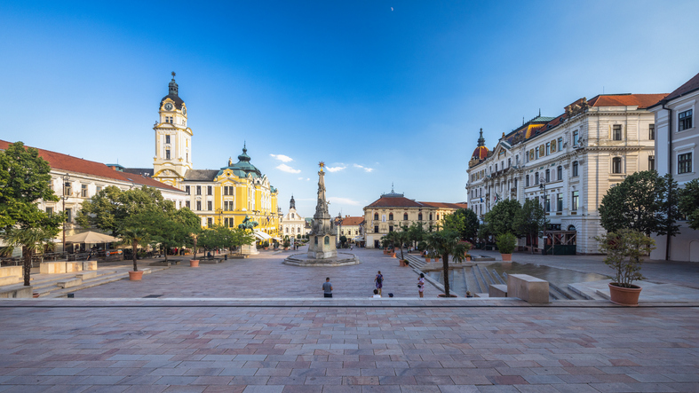 Großer Platz in Pécs mit historischer Kirche, Denkmal, hellen Fassaden und blauem Himmel.