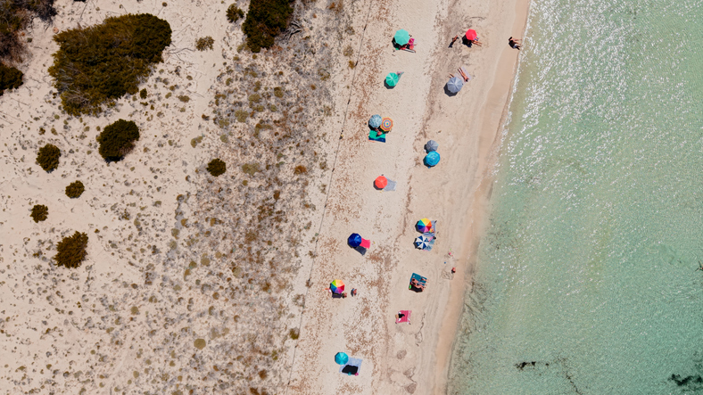 Senkrechte Luftaufnahme eines schmalen Sandstrands von Es Trenc mit bunten Sonnenschirmen am hellen Ufer.