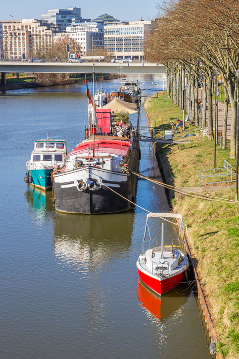 Mehrere Boote liegen am Ufer der Saar in Saarbrücken, daneben verläuft eine Uferpromenade mit Bäumen.