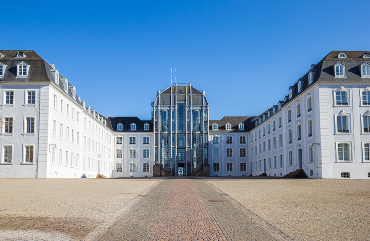 Das Schloss Saarbrücken mit weißer Fassade, weitem Vorplatz und modernem Glaseingang unter blauem Himmel.