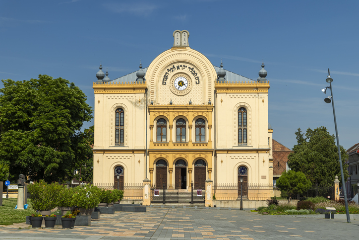Synagoge in Pécs mit markanter gelber Fassade, hebräischer Schrift, Uhr und blauem Himmel.