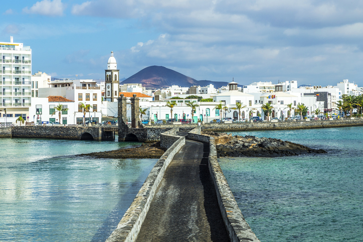 Eine schmale Steinbrücke führt über das Wasser in Richtung Arrecife mit weißen Häusern und Vulkan im Hintergrund.