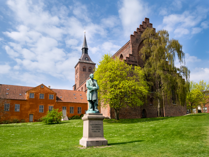 Das Hans-Christian-Andersen-Denkmal steht vor einer Kirche und historischen Gebäuden in Odense.