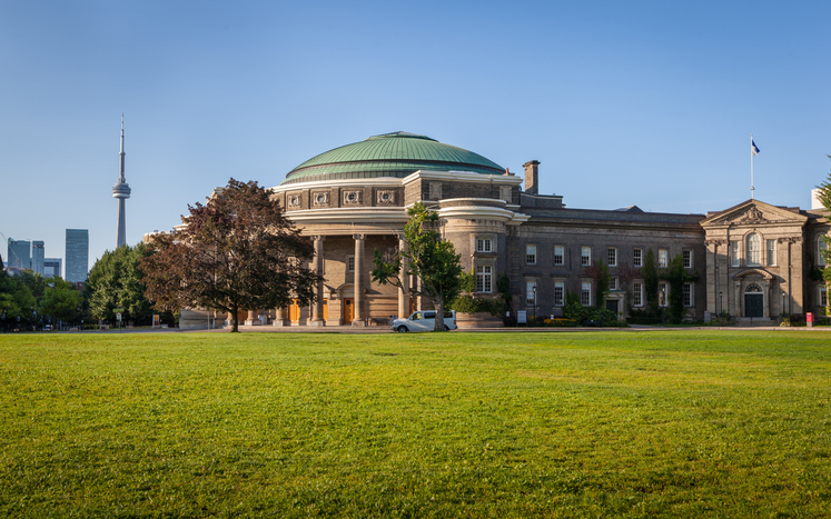 Klassisches Gebäude der University of Toronto mit großer Wiese und CN Tower im Hintergrund.