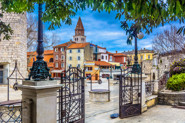Ein geöffneter Torbogen führt auf einen Platz mit Brunnen, Kirchturm und alten Häusern in Zadar.