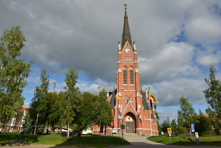 Rote Backsteinkirche mit hohem Turm, umgeben von Bäumen und Grünflächen in Luleå.