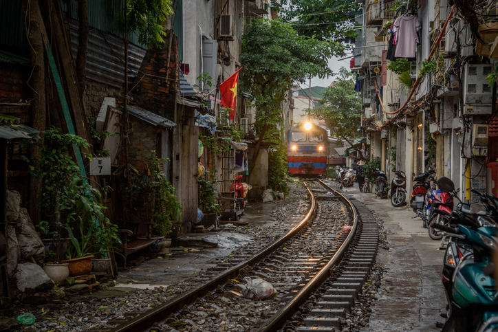 Ein Zug fährt durch eine enge Wohnstraße in Hanoi, die dicht von Häusern und Pflanzen gesäumt ist.