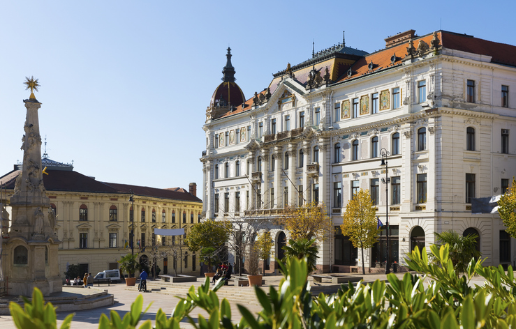 Historisches Gebäude in Pécs mit verzierter Fassade, Platz, Denkmal und sonnigem Stadtbild.
